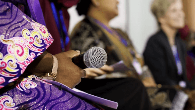 Woman holding a microphone during a panel discussion at a professional women's business event.