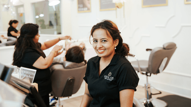 A woman in a black shirt with a logo on it is smiling while sitting at a beauty salon reception desk. In the background, other people are receiving beauty treatments.