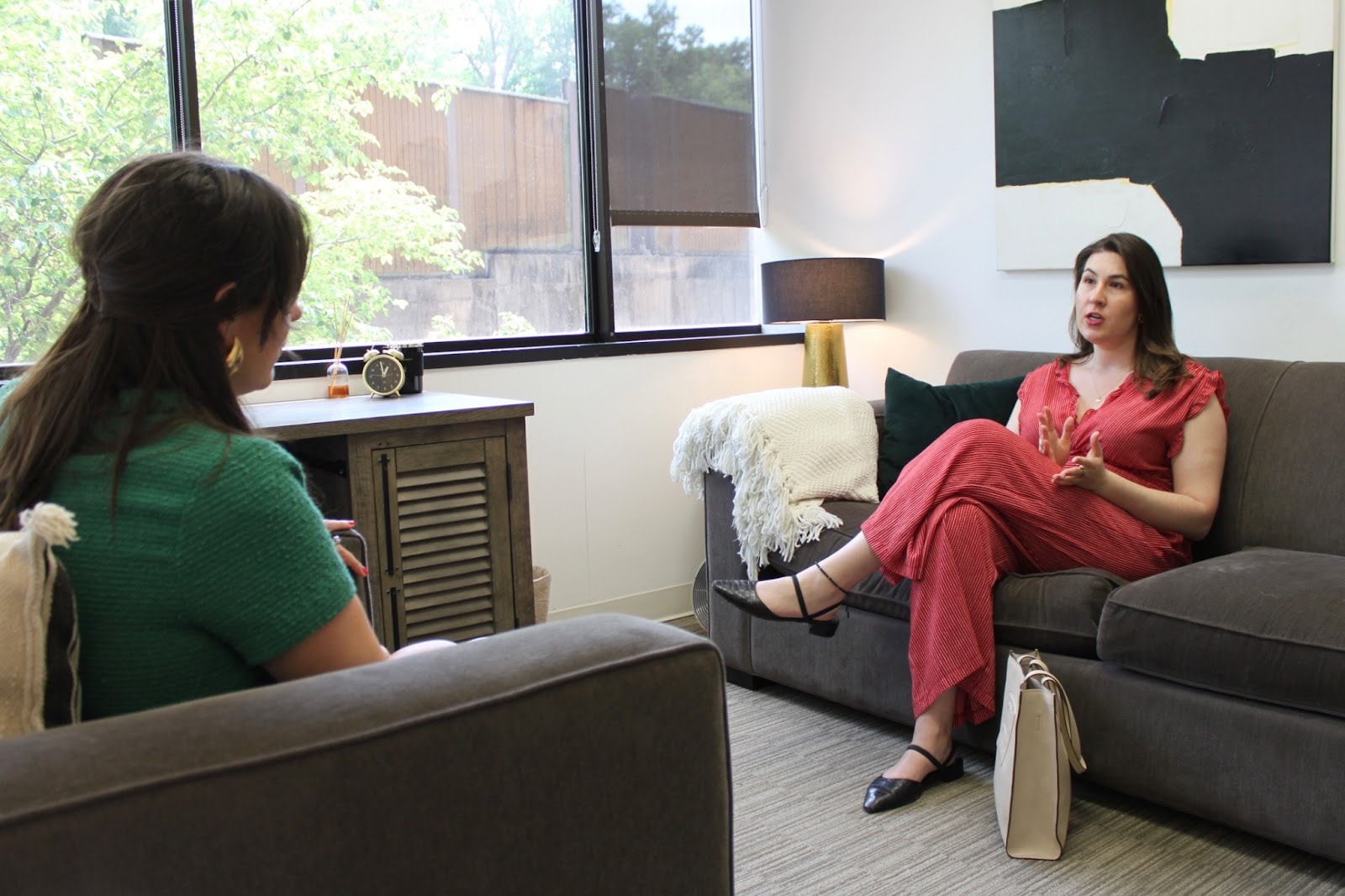Alt text: "Two women engaged in a conversation in a cozy office setting. One woman, wearing a green sweater, is seated on a gray couch with her back facing the camera. The other woman, dressed in a red striped jumpsuit, is sitting on a separate gray couch, gesturing as she speaks. A wooden cabinet, table lamp, and window with greenery outside are visible in the background.