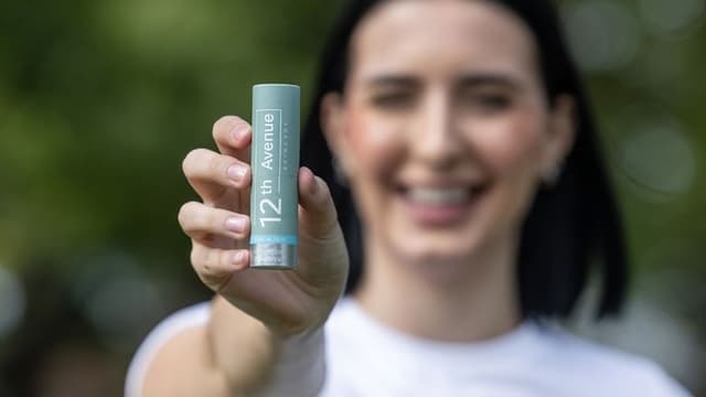 A smiling woman holding the 12th Avenue skincare HydraRetin balm stick in front of a blurred natural background, promoting its dual-action formula for hydration and skin care."